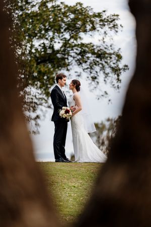 Bride and groom stand together under a tree framed by branches, bride holding a bouquet on their wedding day Indooroopilly Golf Club QLD