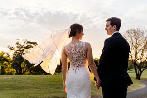 Bride and groom holding hands at sunset, her lace gown and veil flowing in the breeze on a lush green lawn at Indooroopilly Golf Club