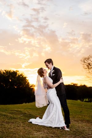 Bride and groom share a romantic kiss at sunset on a green lawn, with golden clouds glowing in the background at Indooroopilly Golf Club