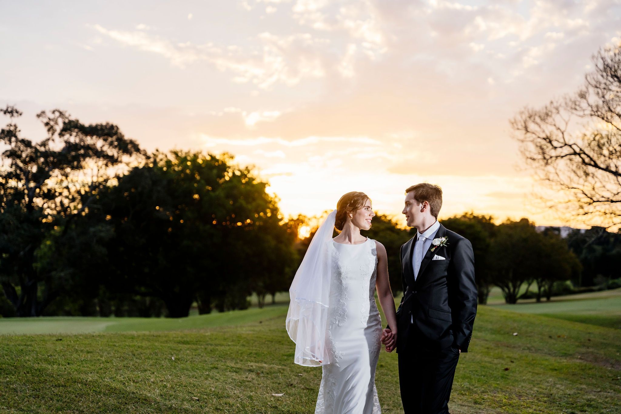 Bride and groom hold hands at sunset on a lush green golf course, golden light glowing behind the trees at Indooroopilly Golf Club Brisbane