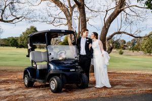 Bride and groom posing beside a blue golf cart on a sunny golf course, surrounded by trees at Indooroopilly Golf Club Brisbane Queensland