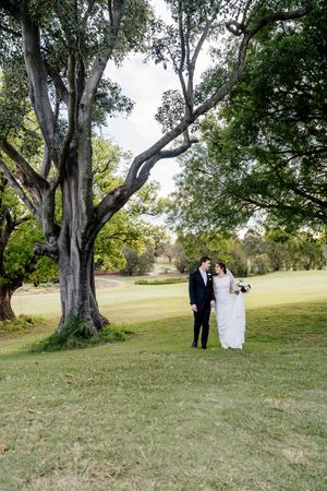 Bride and groom walk hand in hand on lush green golf course grounds beneath sprawling trees on their wedding day Indooroopilly Golf Club