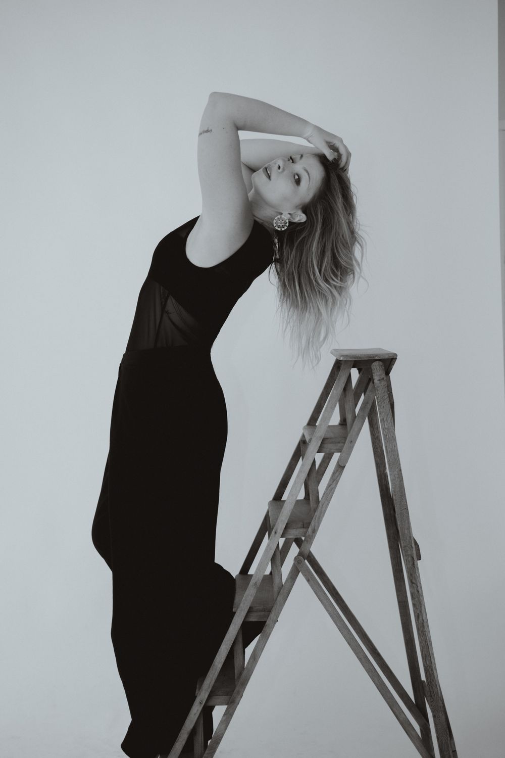Black and white photo of a woman in a black dress posing gracefully beside a wooden stepladder.