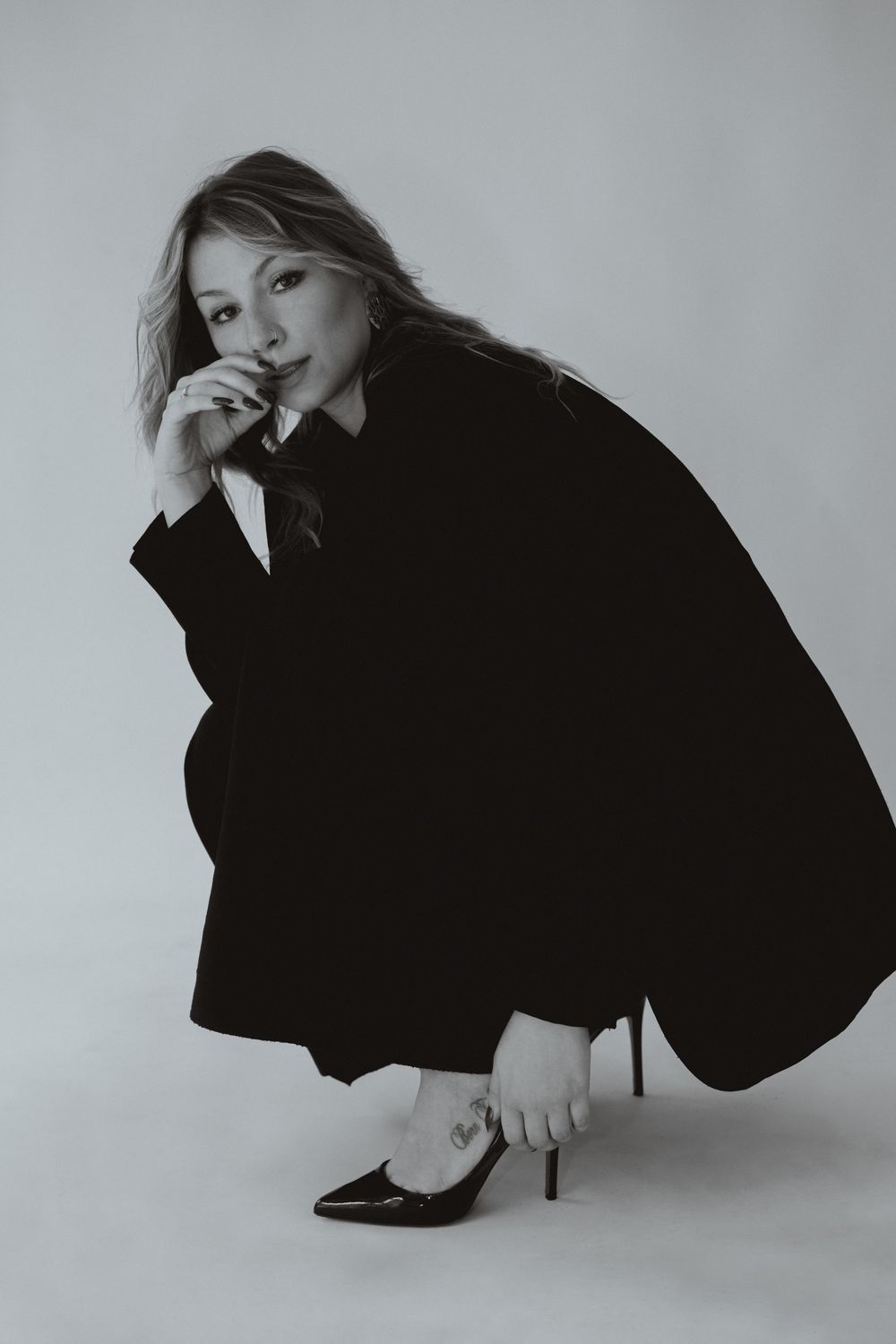 Black and white photo of a woman in an oversized black blazer and heels, sitting gracefully on a studio floor.