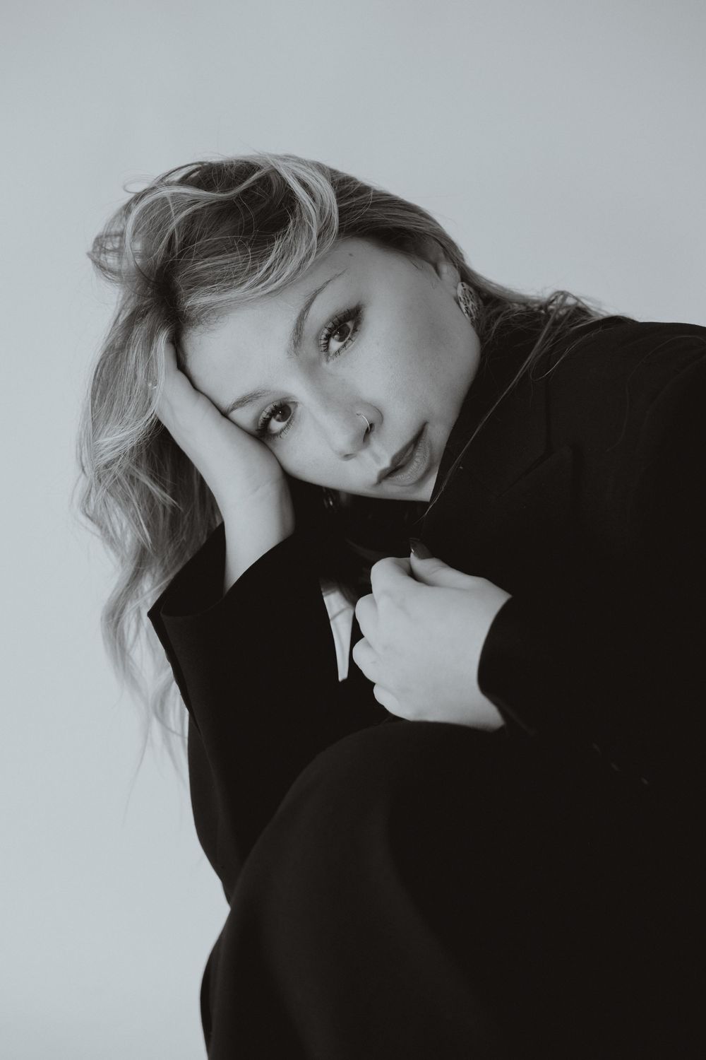 Black and white portrait of a young blonde woman in a dark coat, resting her head on her hand, gazing softly at camera.