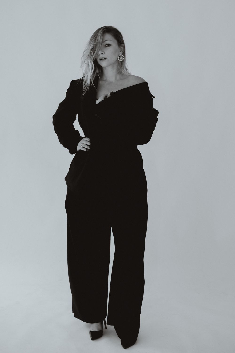 Woman in black off-shoulder jumpsuit and heels poses confidently in a black and white studio photo.