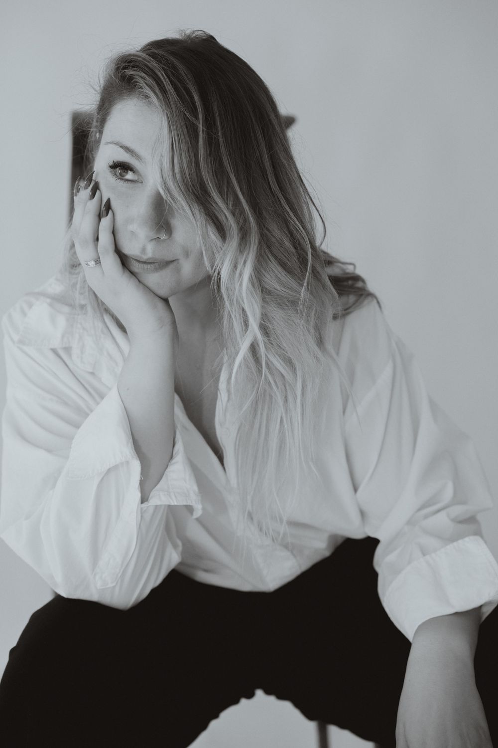 Black and white portrait of a blonde woman in a white shirt resting her chin on her hand, gazing thoughtfully upward.
