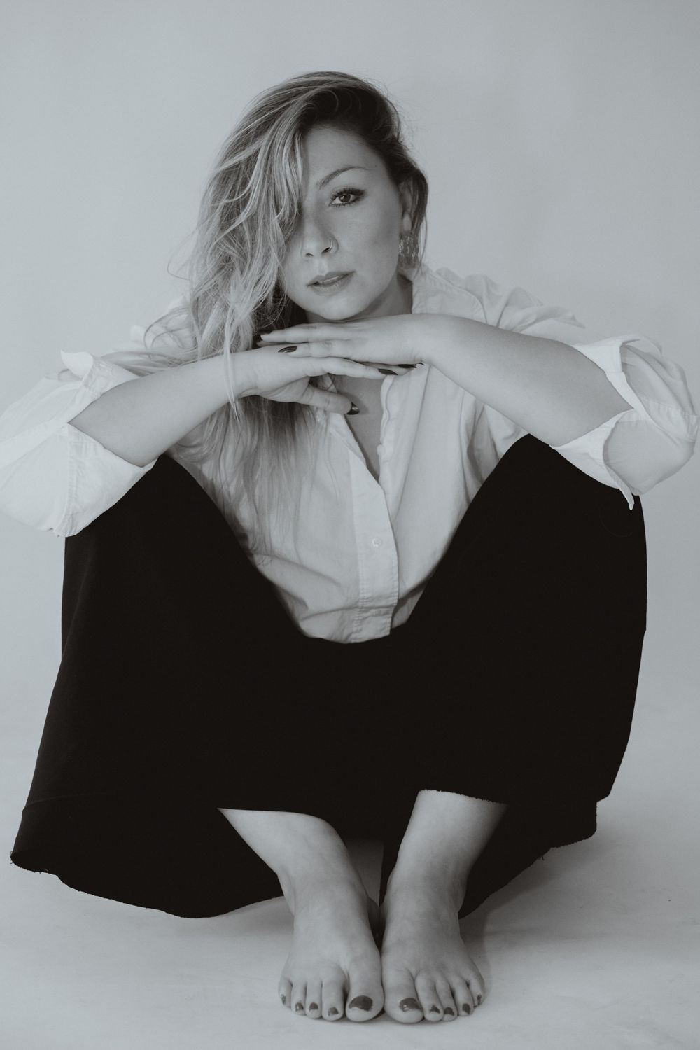 Black and white portrait of a blonde woman sitting barefoot on floor in white shirt and black pants, resting chin on hands.