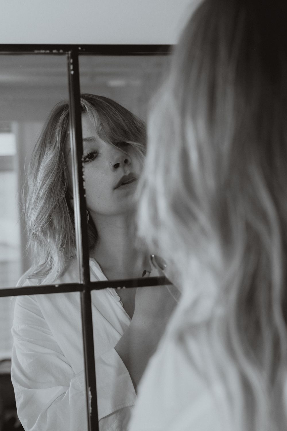 Black and white photo of a blonde woman in a white shirt gazing at her reflection in a framed mirror.