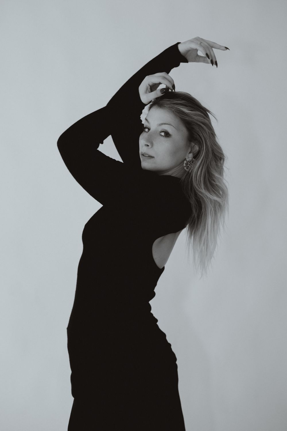 Black and white portrait of a blonde woman in a black long-sleeve dress posing gracefully with one arm raised.