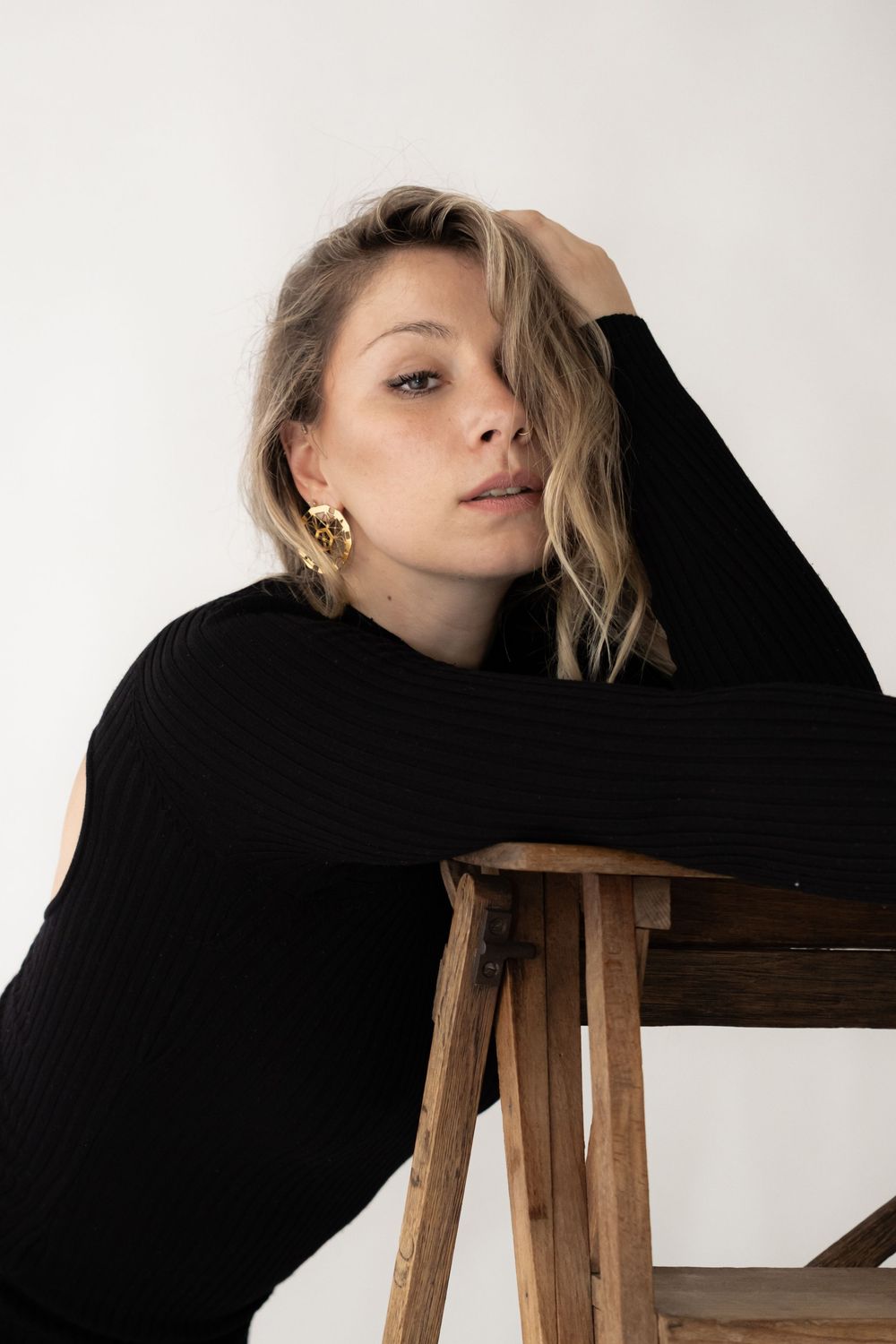 Woman in black ribbed sweater leaning on wooden stool, wearing gold earrings, with blonde hair against white background.