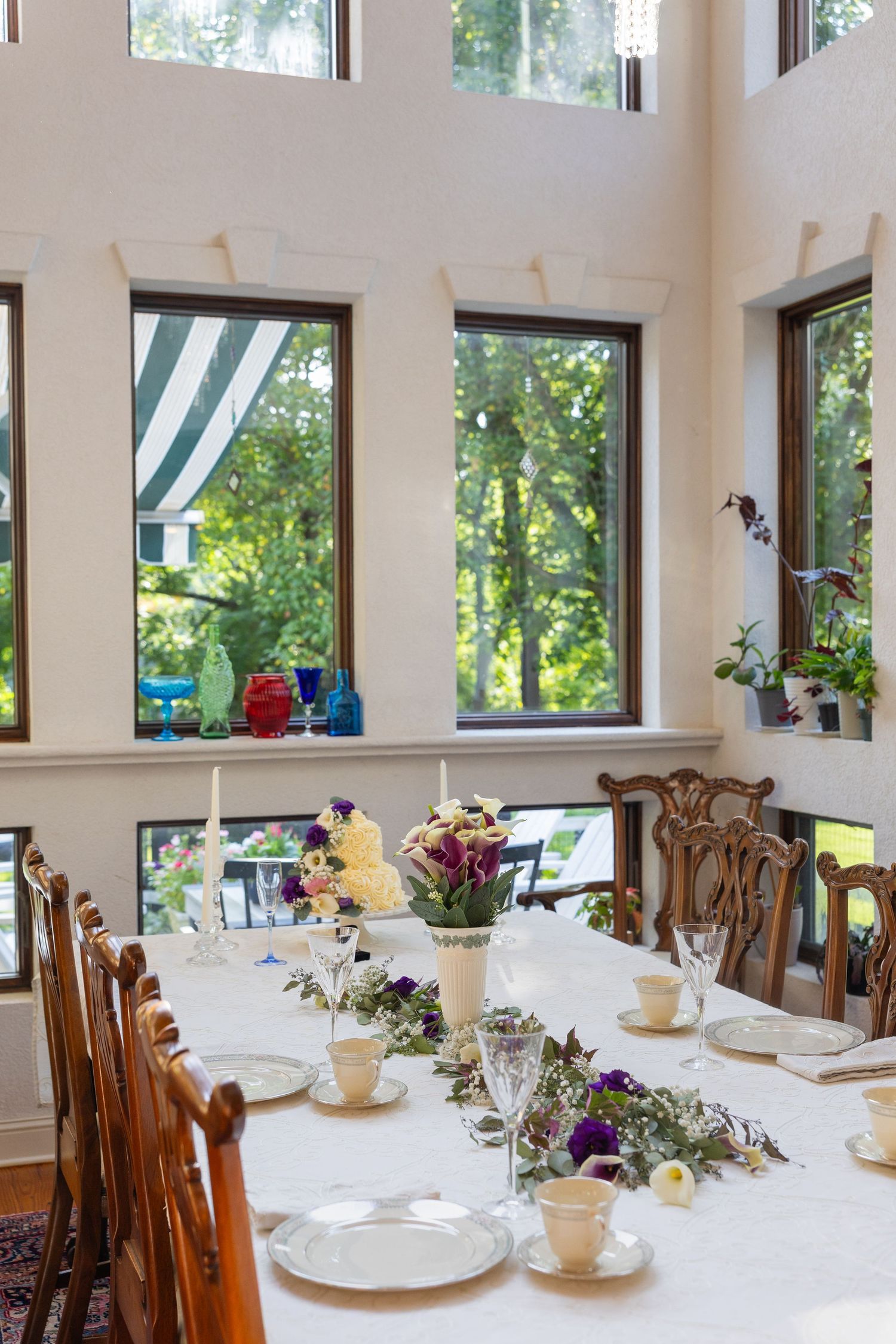 Elegant bed and breakfast interior with floor-to-ceiling windows, set dining tables, and a brick fireplace surrounded by lush greenery.