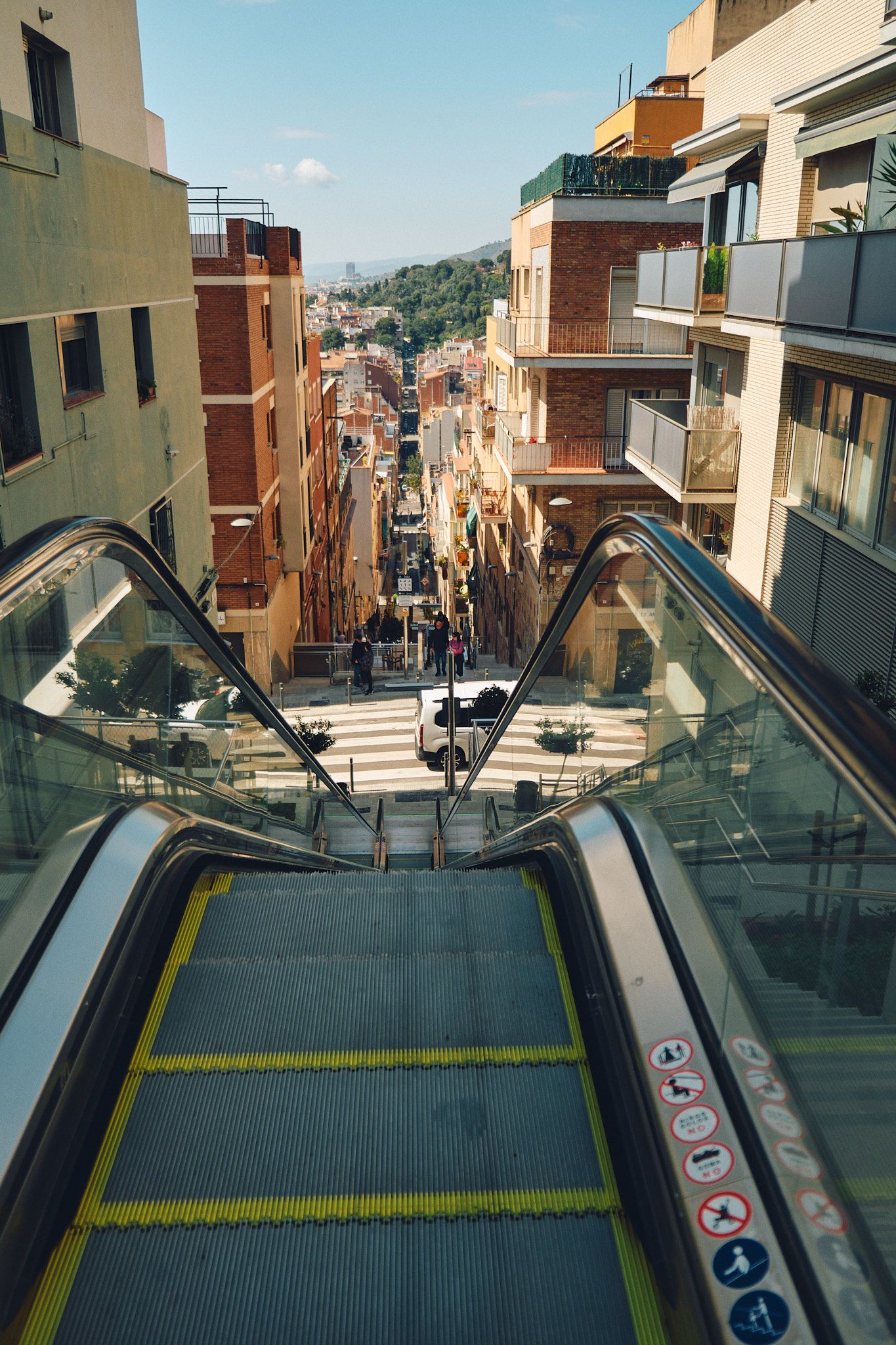 Outdoor escalators descending a steep urban street flanked by modern apartment buildings in Barcelona, Spain.