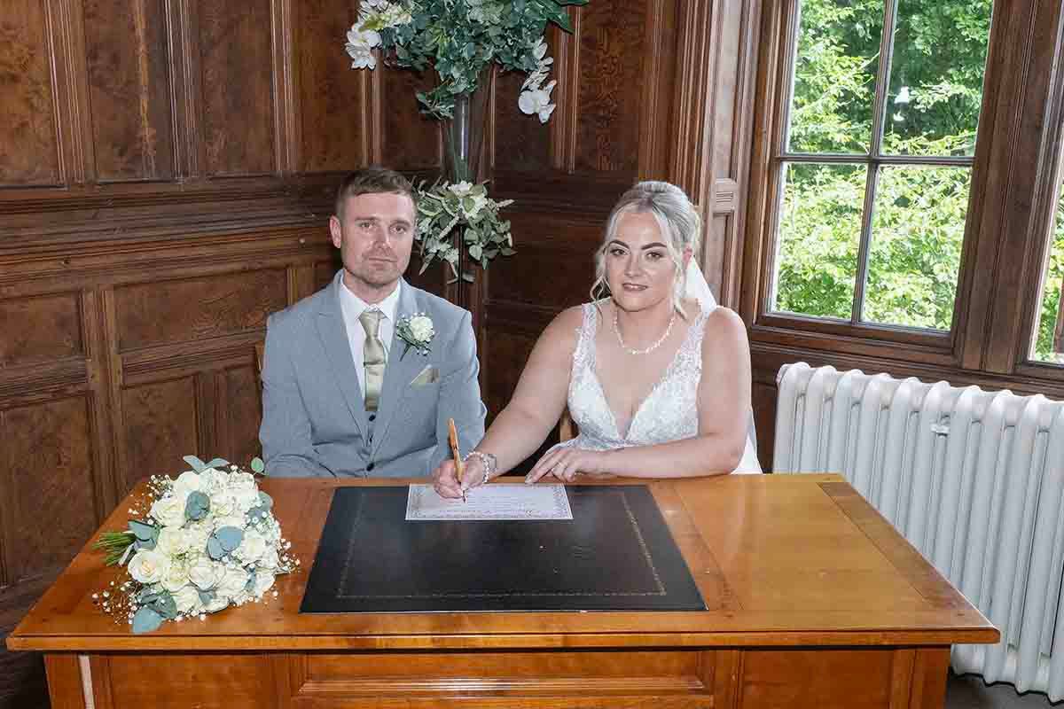 Bride signing wedding register at wooden desk beside groom in grey suit in elegant wood-panelled registry office.