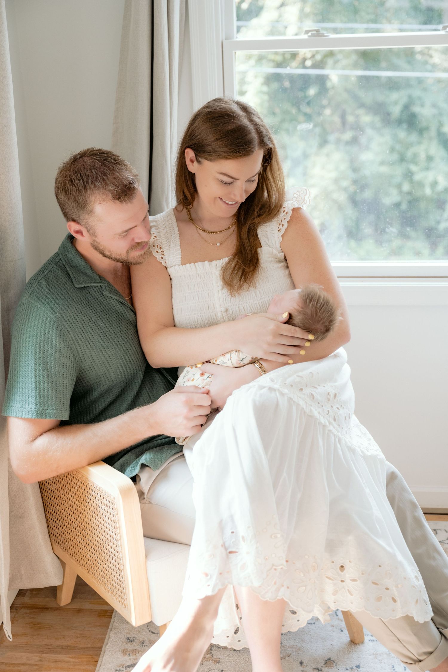 A young mom and dad sit together in a well lit living space cradling their brand new baby girl during an in-home lifestyle newborn session.