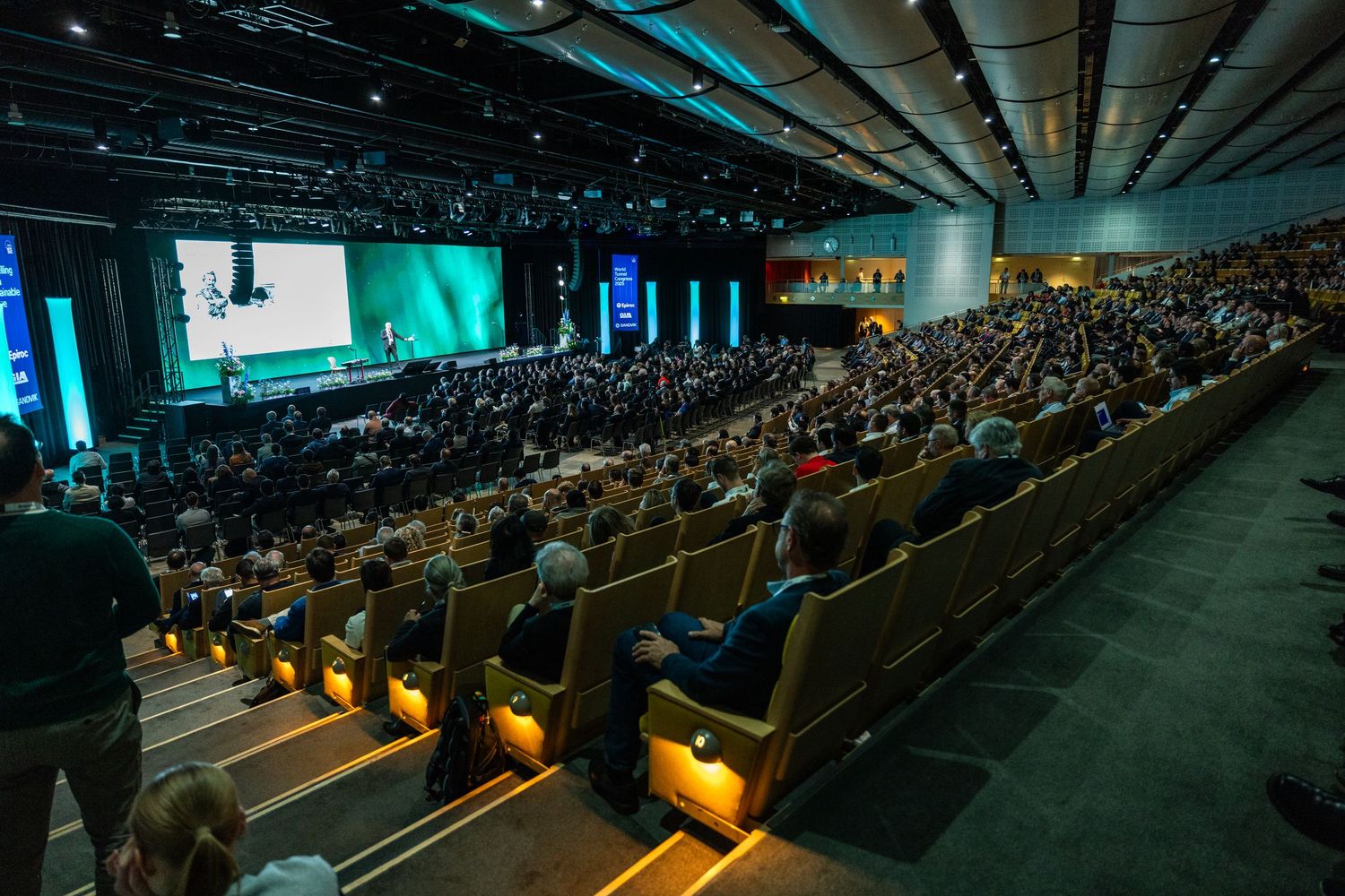 A large auditorium filled with hundreds of attendees watching a speaker on a green-lit stage.