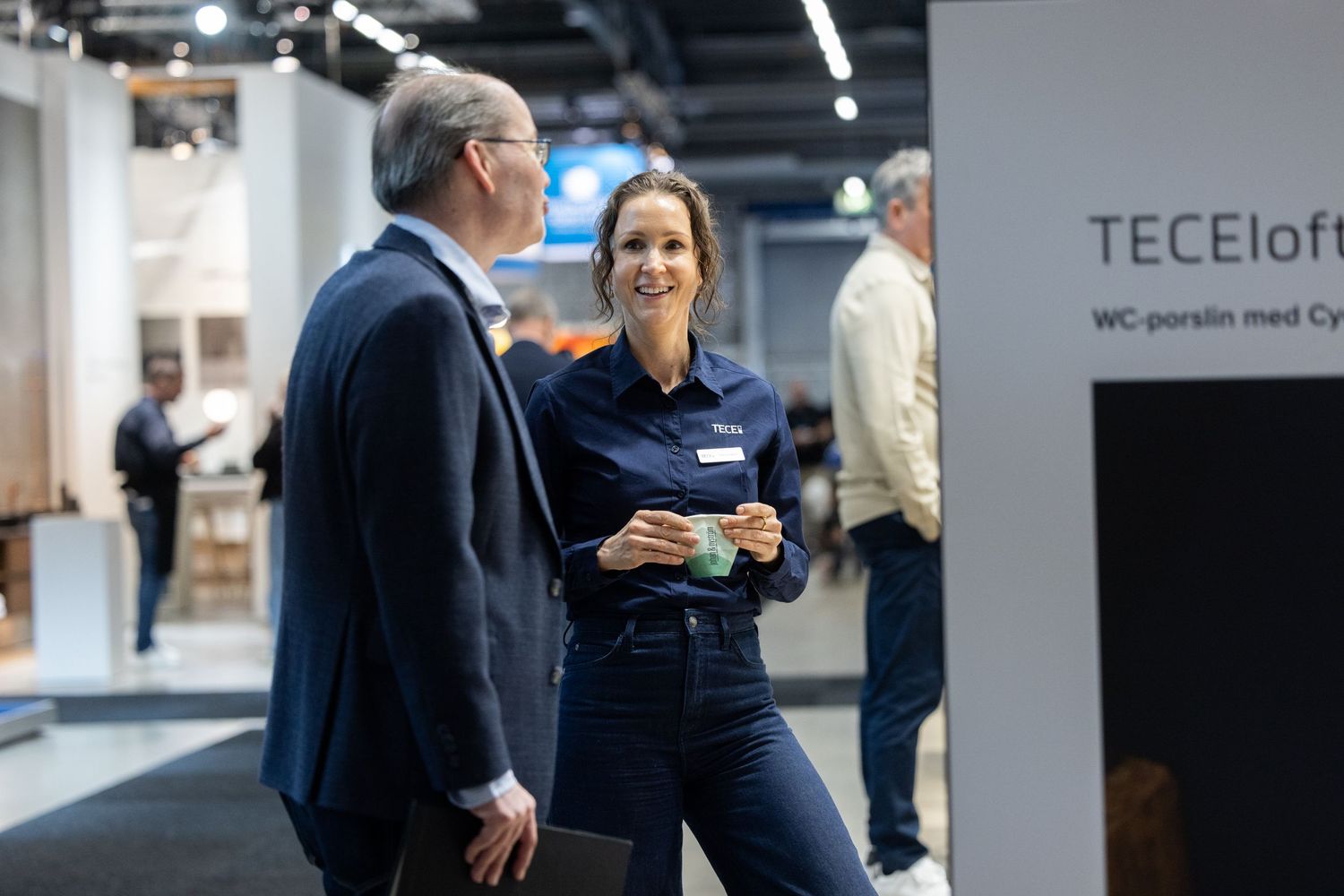 Business professionals engage in conversation near a TECEloform display at a trade show.