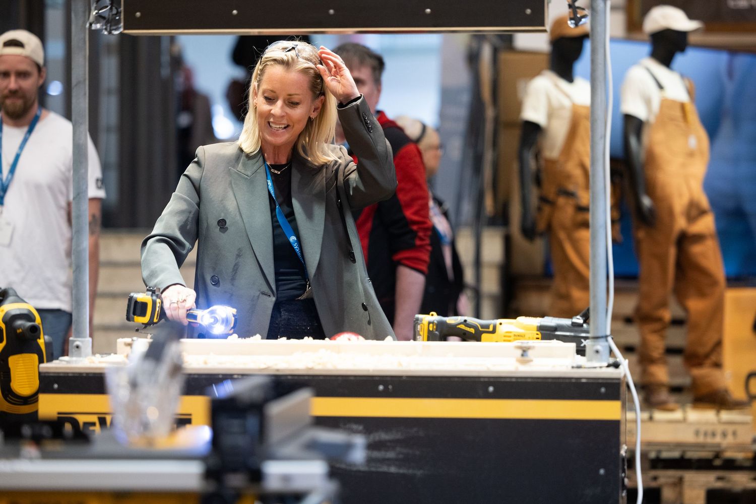 Woman in blazer demonstrates power tools at a busy trade show exhibition stand.