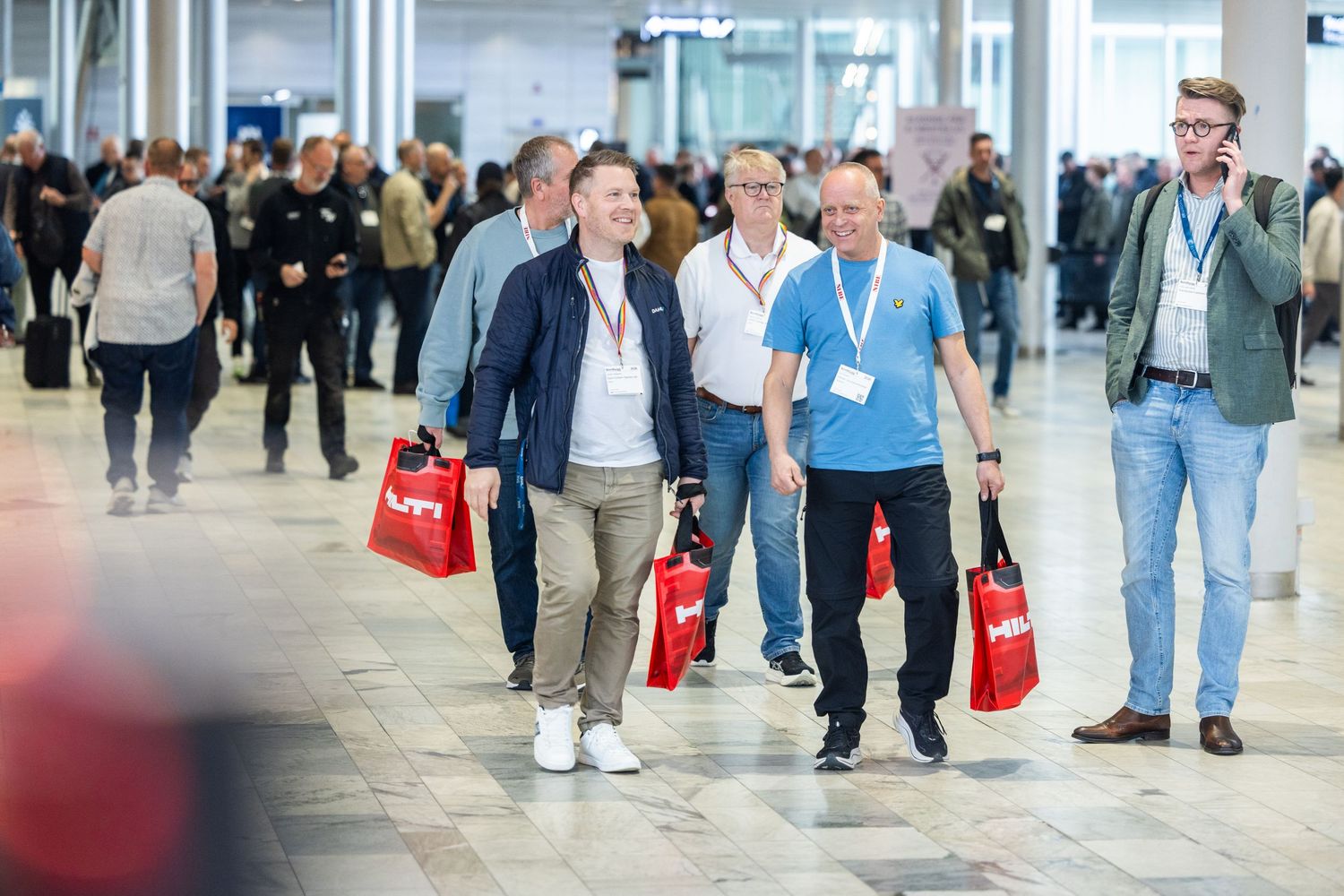Business attendees walk through a convention center carrying red shopping bags at a trade fair.