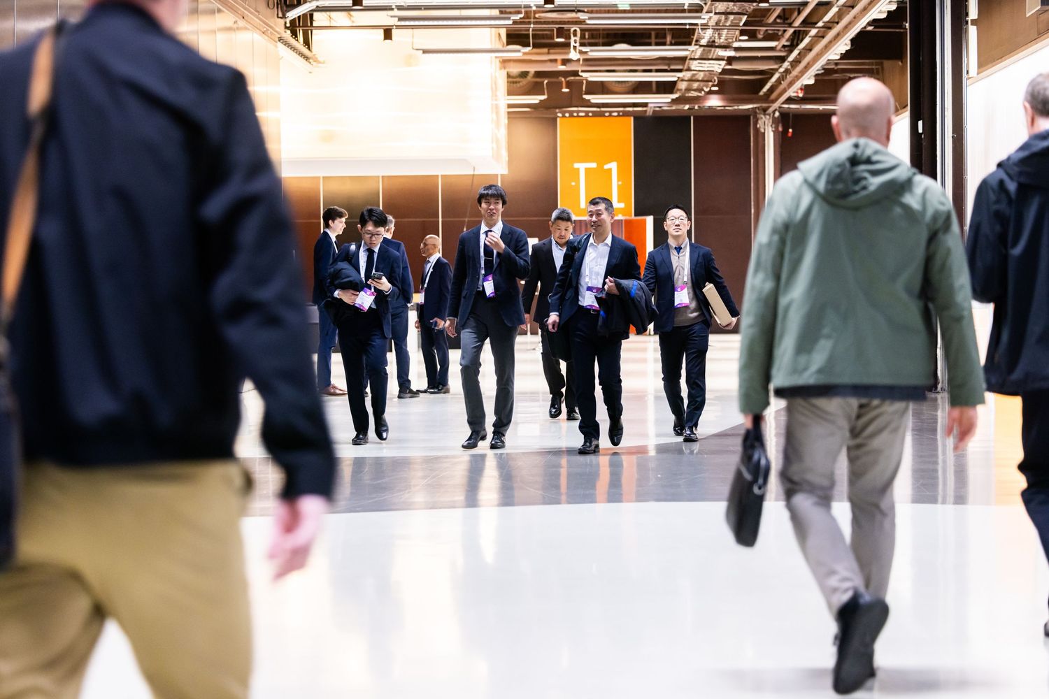 A group of professionals in suits walk purposefully through a modern convention center corridor.