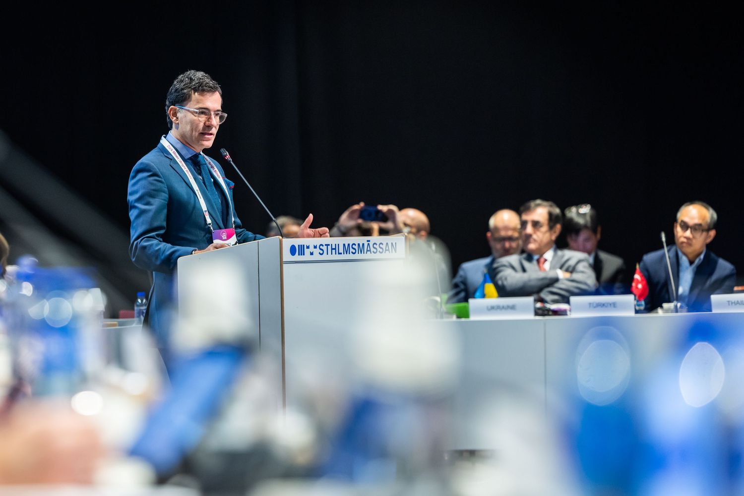 A man in a blue suit delivers a speech at a podium during a formal conference session.