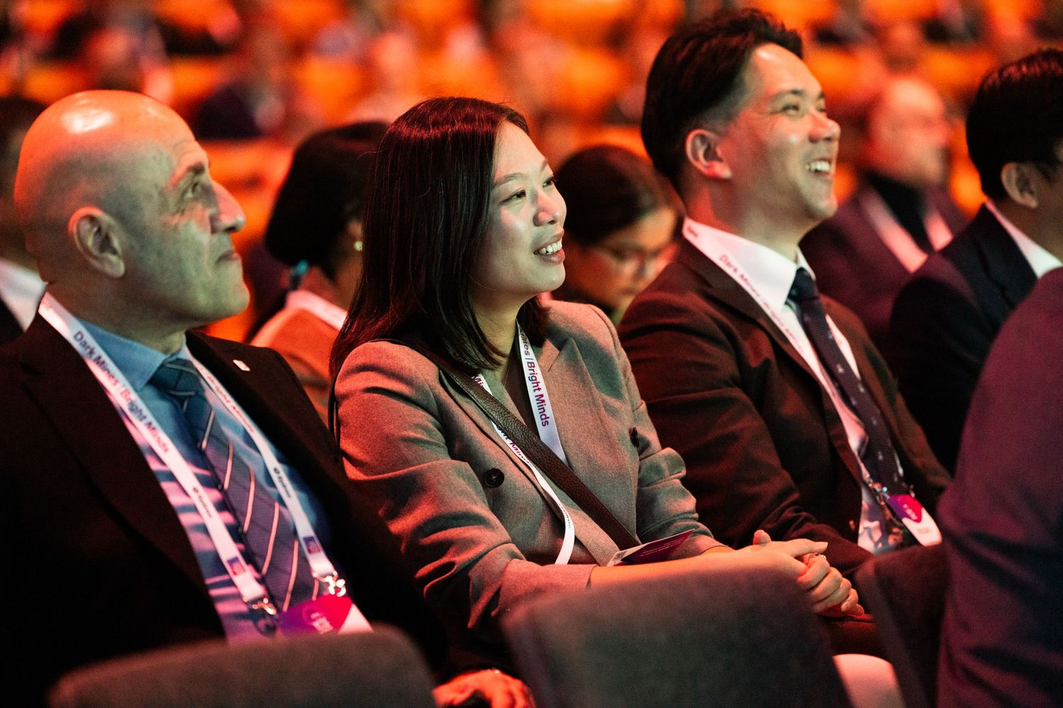 Three conference attendees smile and laugh together while seated in an auditorium with red lighting.