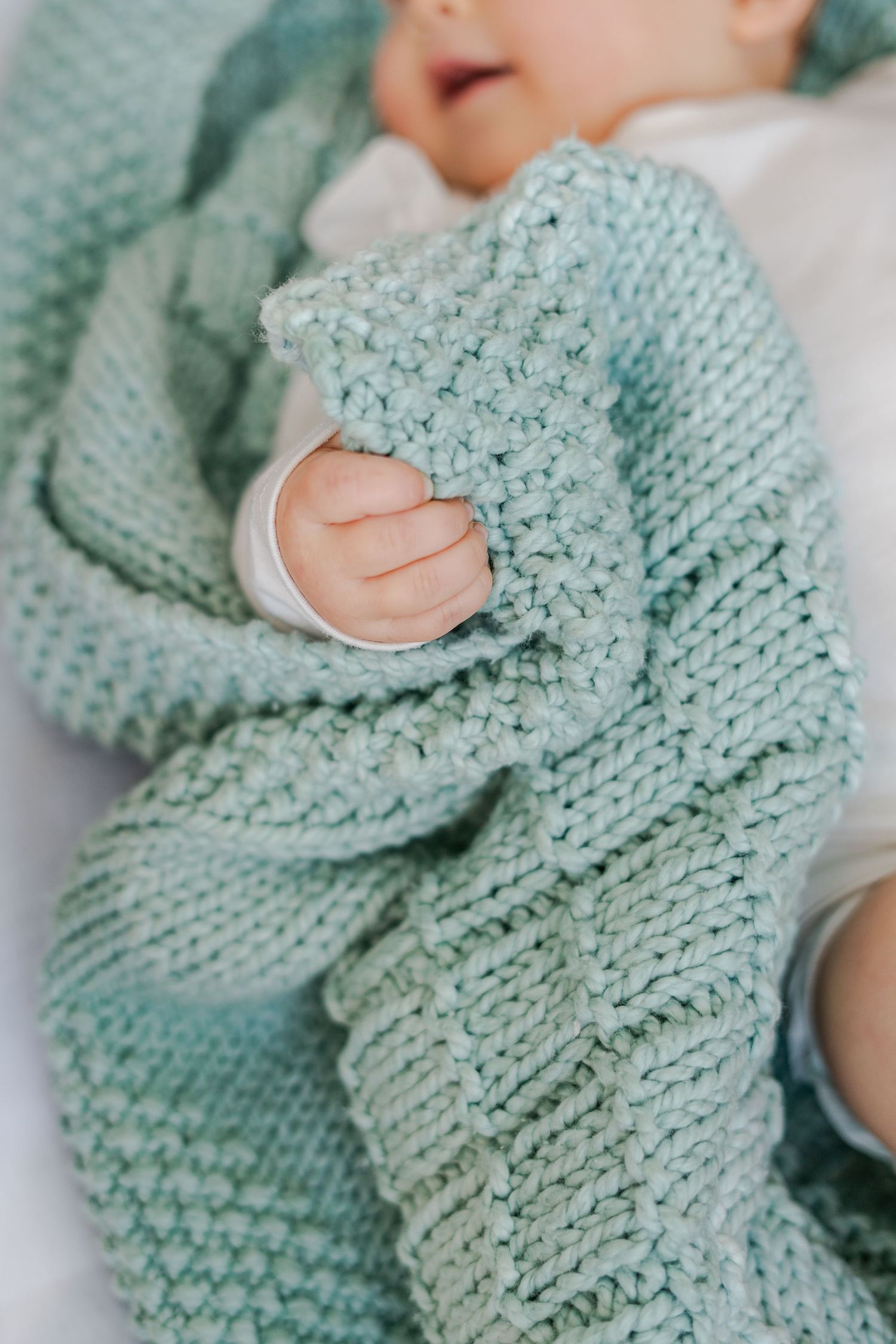 Detail product photography of a baby wrapped in a soft mint green knitted blanket, gently clutching the cozy texture with tiny fingers.