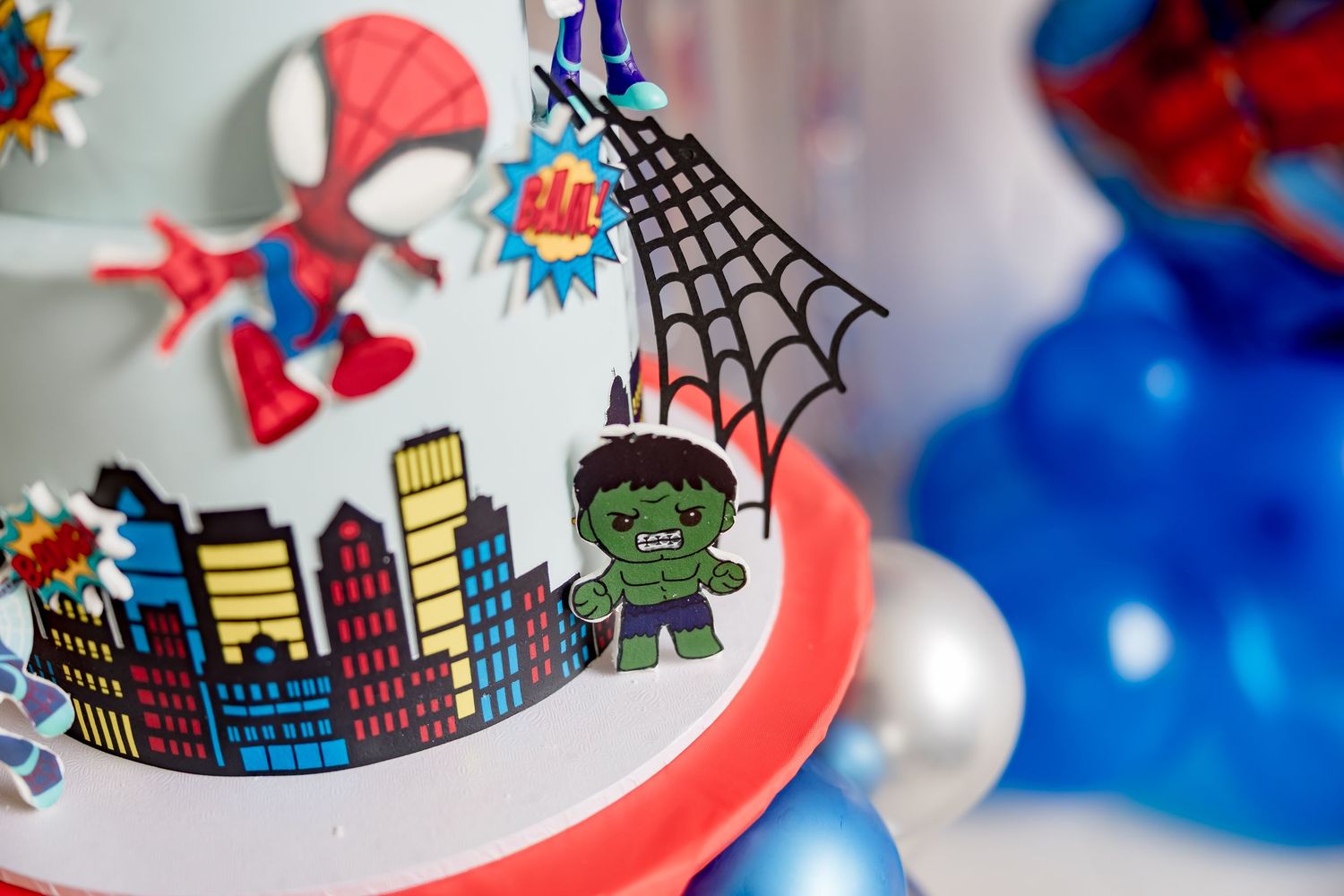 Superhero birthday cake featuring Hulk and Spider-Man decorations with colorful cityscape design and spider web topper.