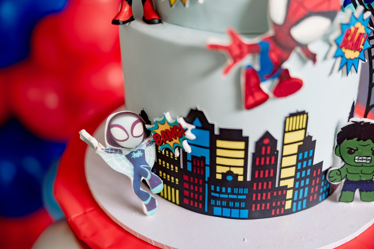 Spider-Man themed birthday cake with Hulk and Ghost-Spider figurines and city skyline.
