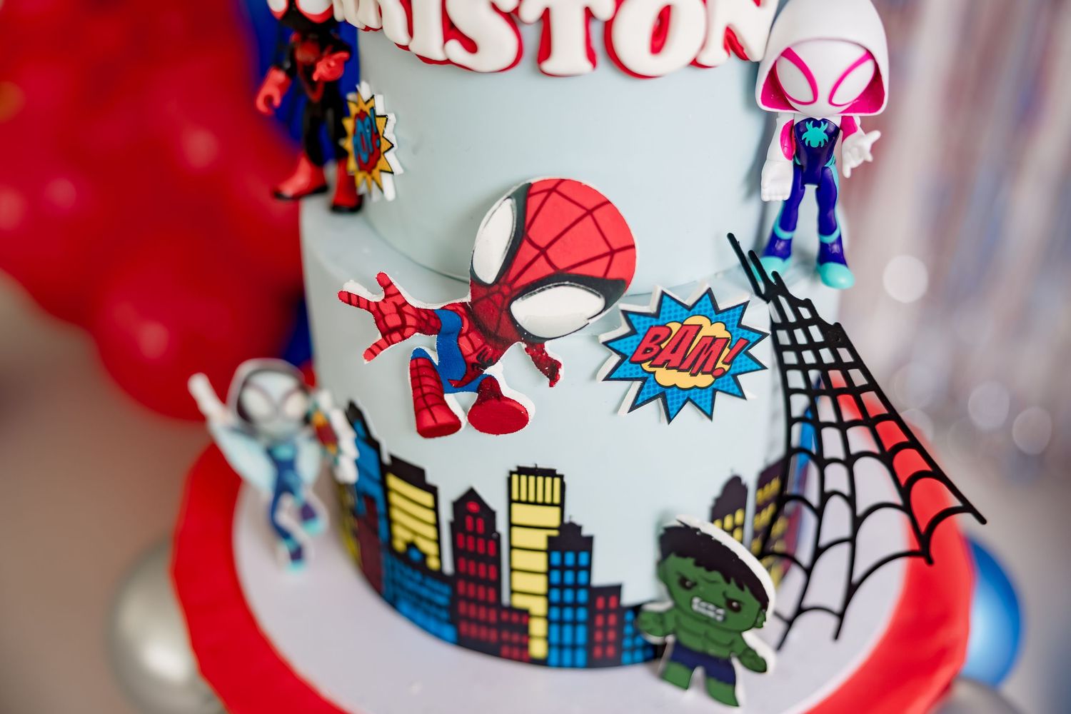 Spiderman themed birthday cake with colorful superhero figurines, city skyline decorations, and BAM comic book text for a child named Aston.