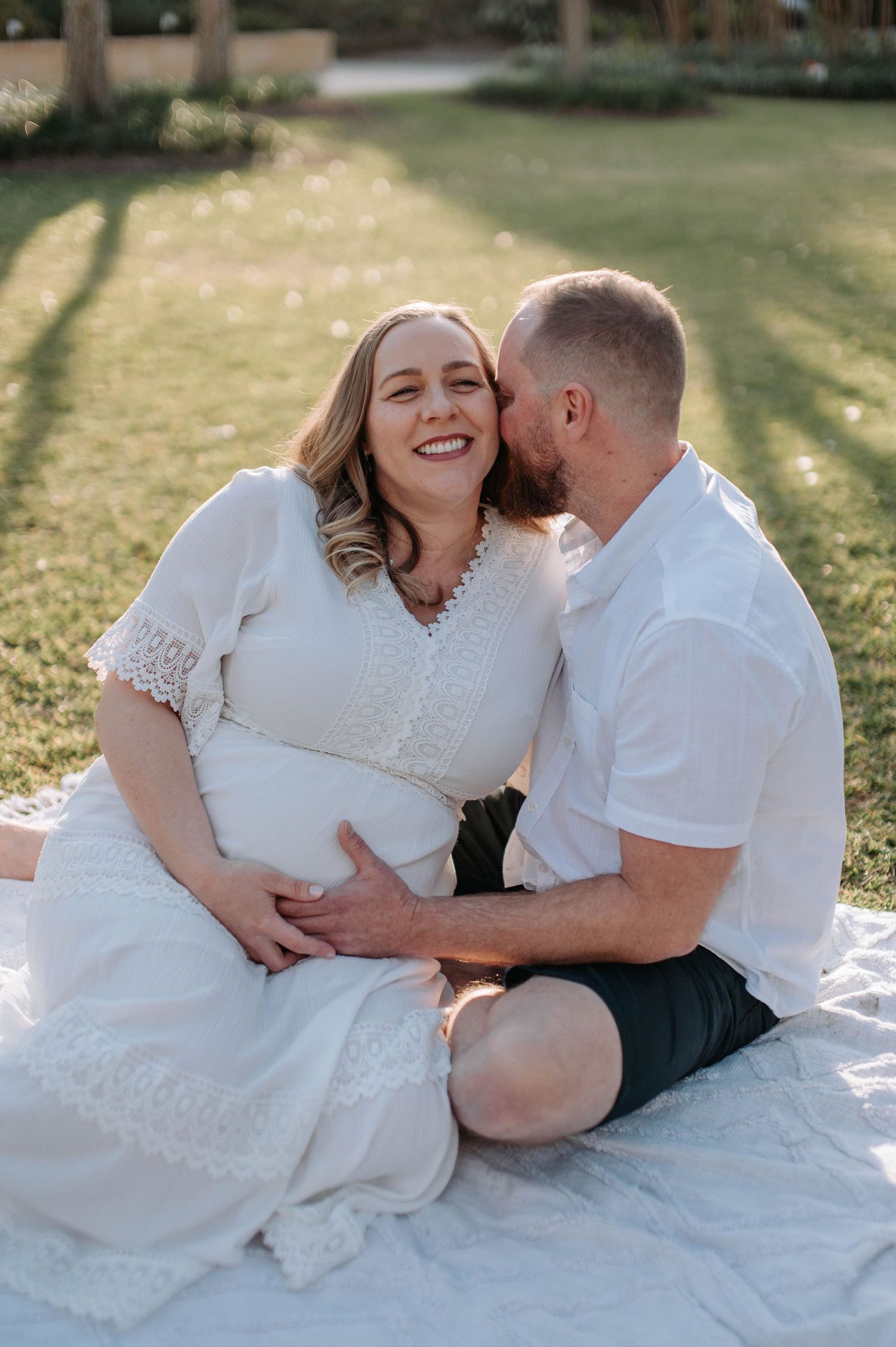 Pregnant woman in white dress smiles as man kisses her cheek while sitting on blanket outdoors.