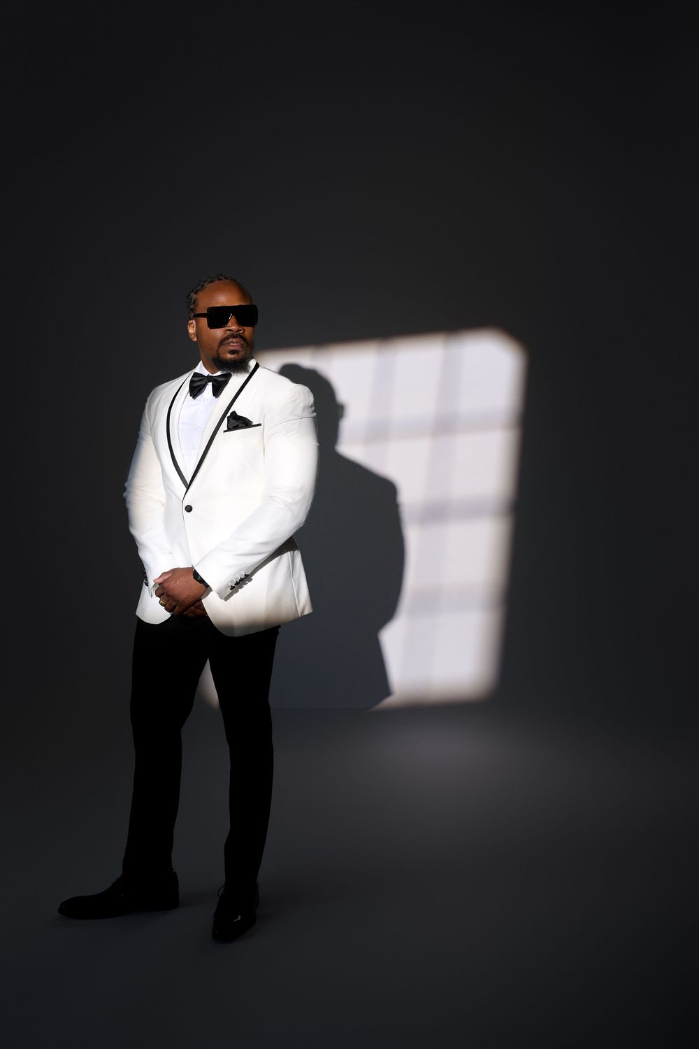 Man in white tuxedo jacket, bow tie, and sunglasses posing confidently in dramatic studio lighting.