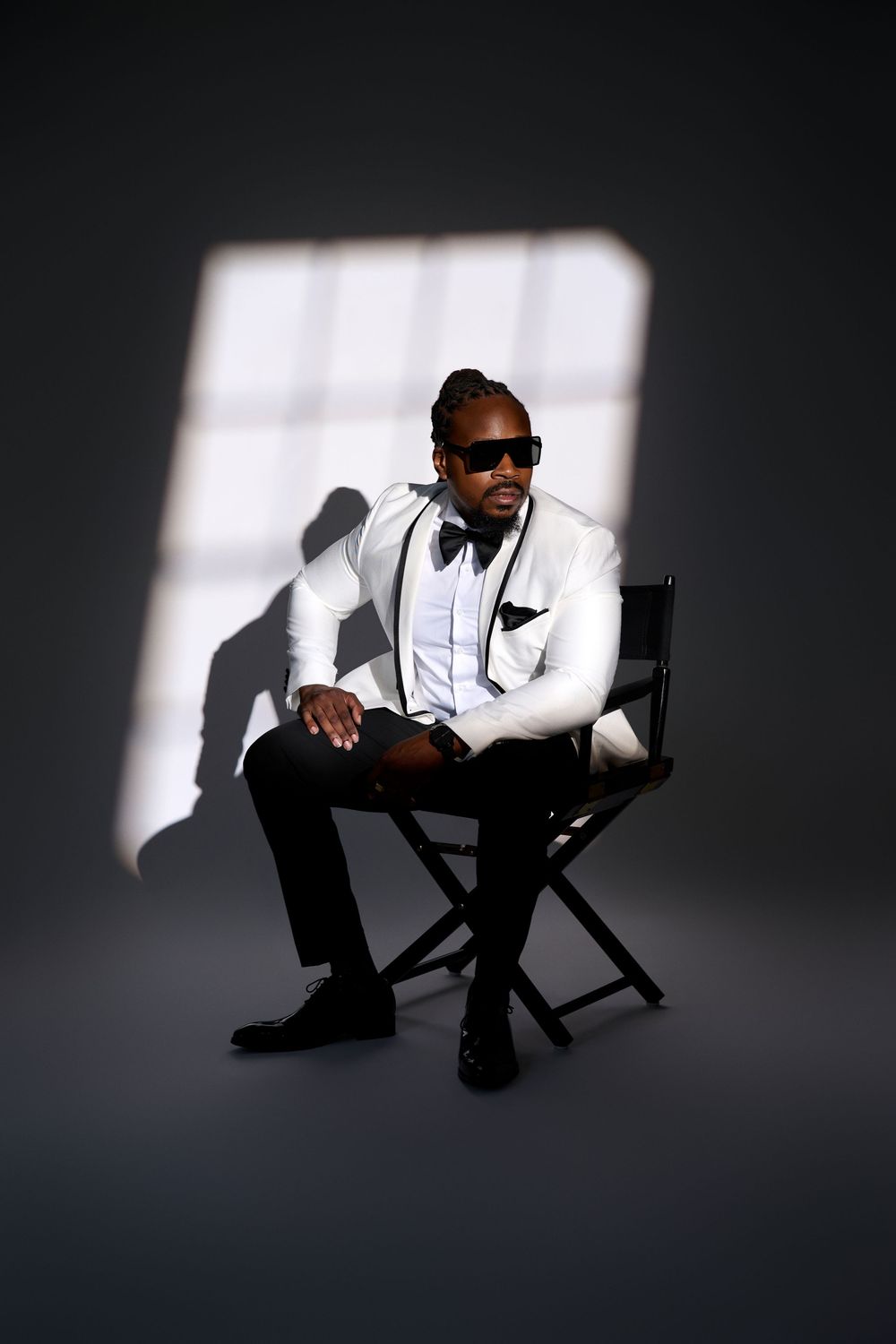 A stylish man in a white tuxedo jacket and sunglasses sits in a director's chair under dramatic studio lighting.