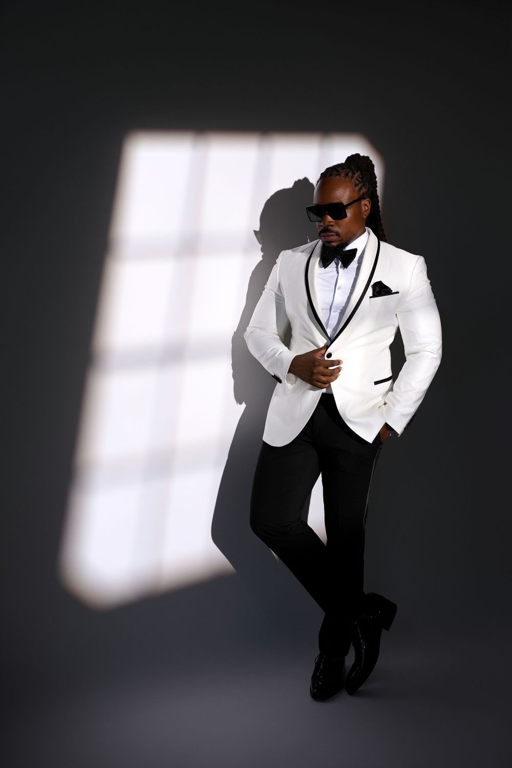 Stylish man in white tuxedo jacket, black pants, and sunglasses posing against dark wall with dramatic light.