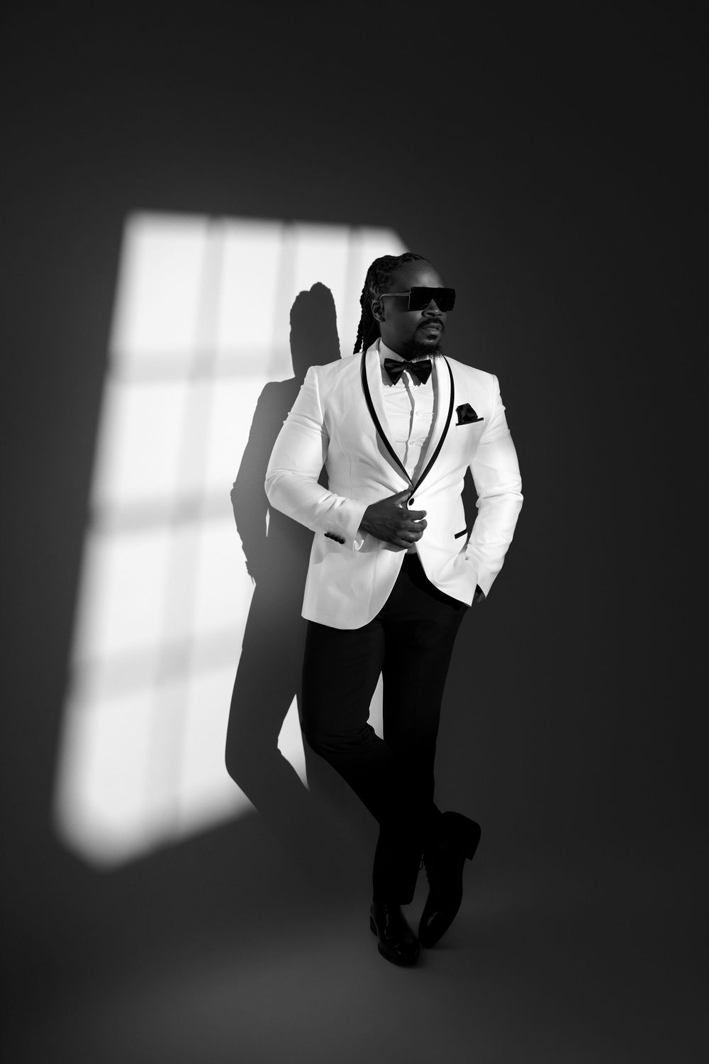 Black and white portrait of a stylish man in white tuxedo jacket, bow tie, and sunglasses leaning against a wall.