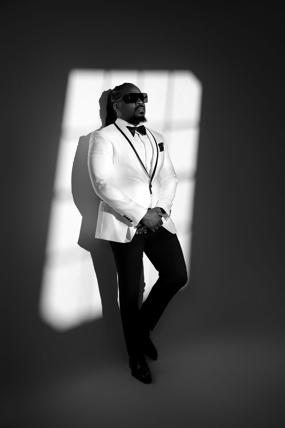 Black and white portrait of a stylish man in white tuxedo jacket, bow tie, and sunglasses posing in dramatic light.