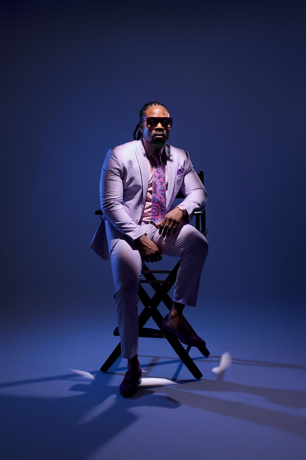 Stylish man in lavender suit and sunglasses sitting confidently in director's chair against blue background.