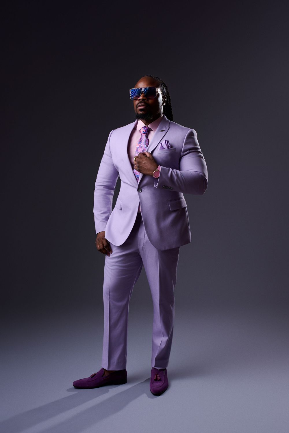 Confident man in lavender suit with pink tie and blue sunglasses posing on gray background.