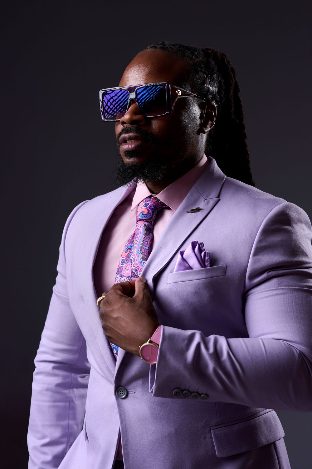 Stylish man in lavender suit, floral tie, pocket square, sunglasses, and braids poses confidently against dark background.