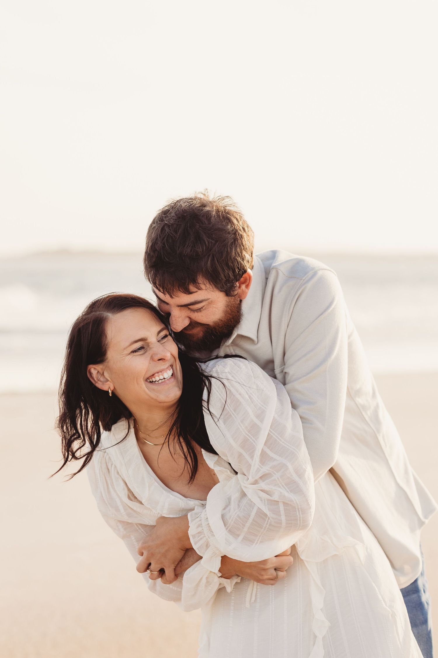 A couple embraces on a beach, the bearded man kissing the laughing woman in a white dress.
