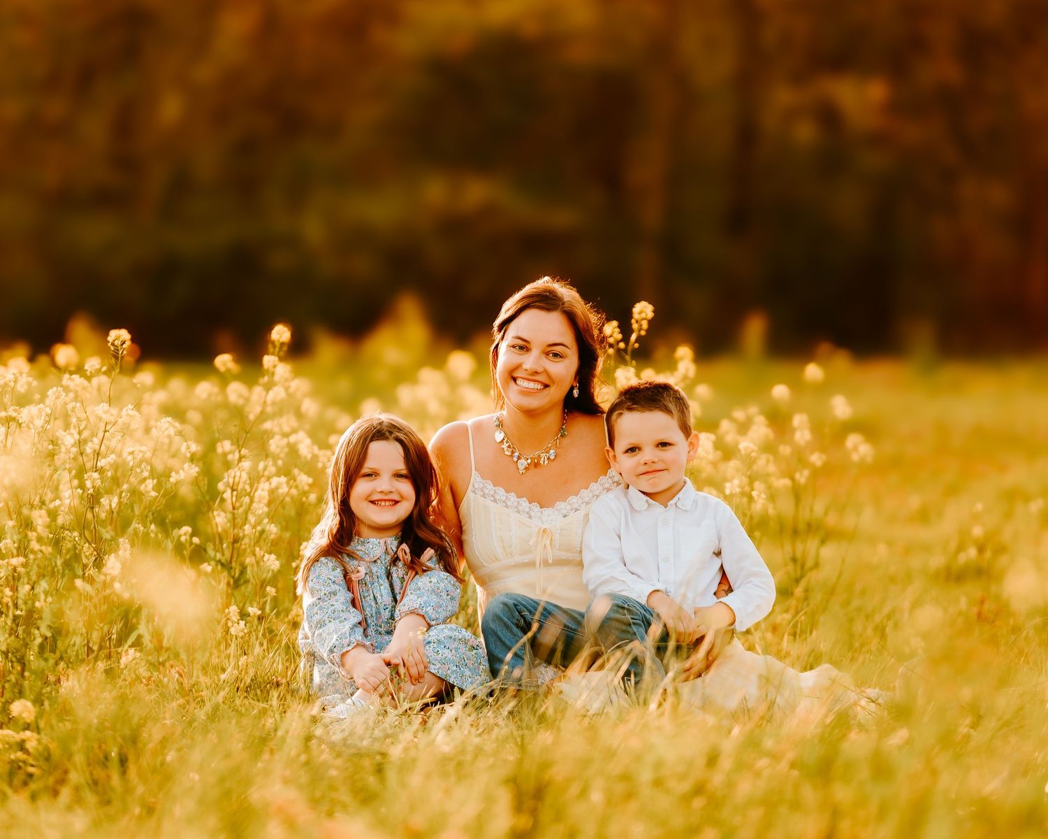 Mother sitting in golden wildflower field with two young children, warm sunset photography.