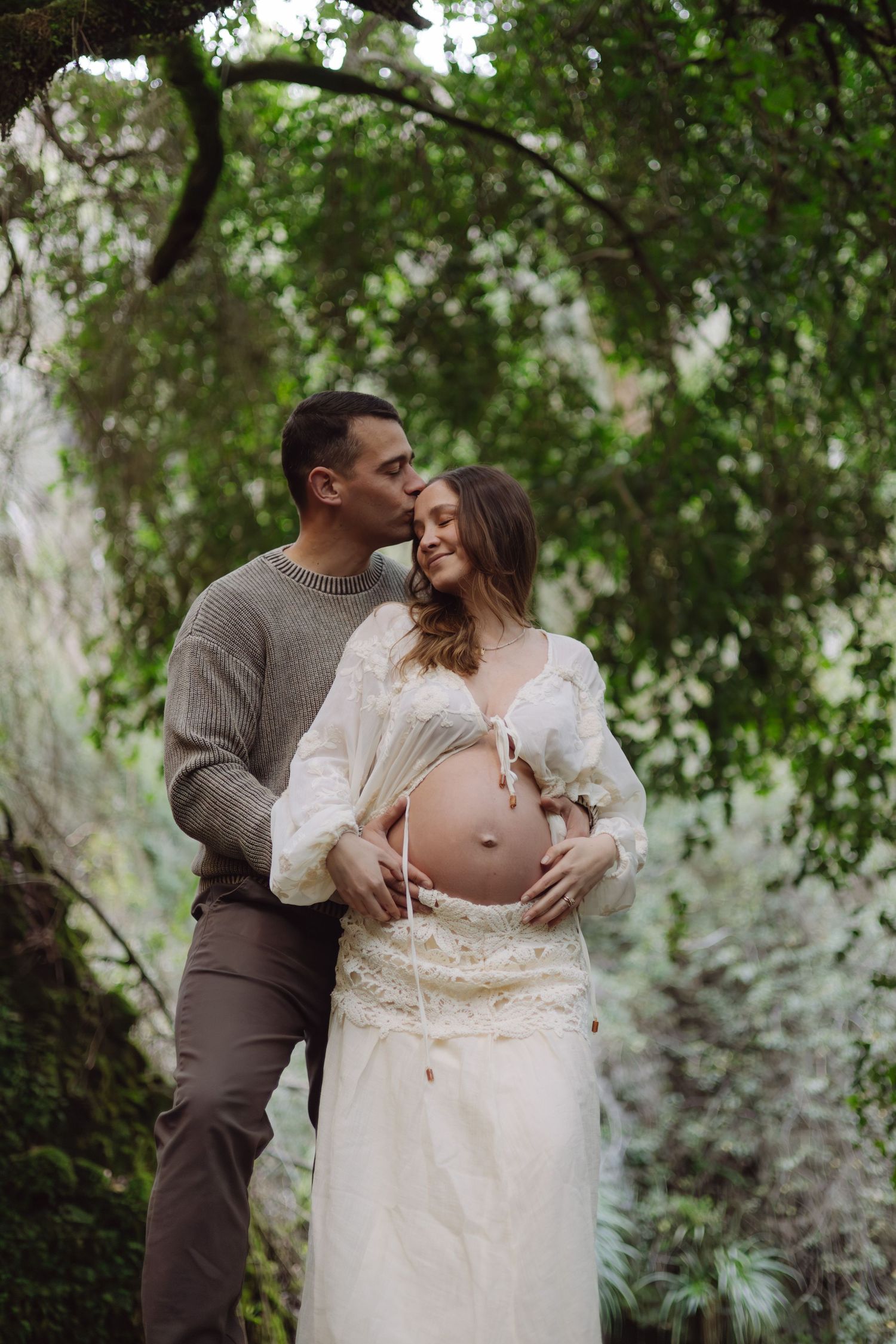 A couple poses outdoors, man kissing pregnant woman's temple while she smiles, surrounded by lush green trees.