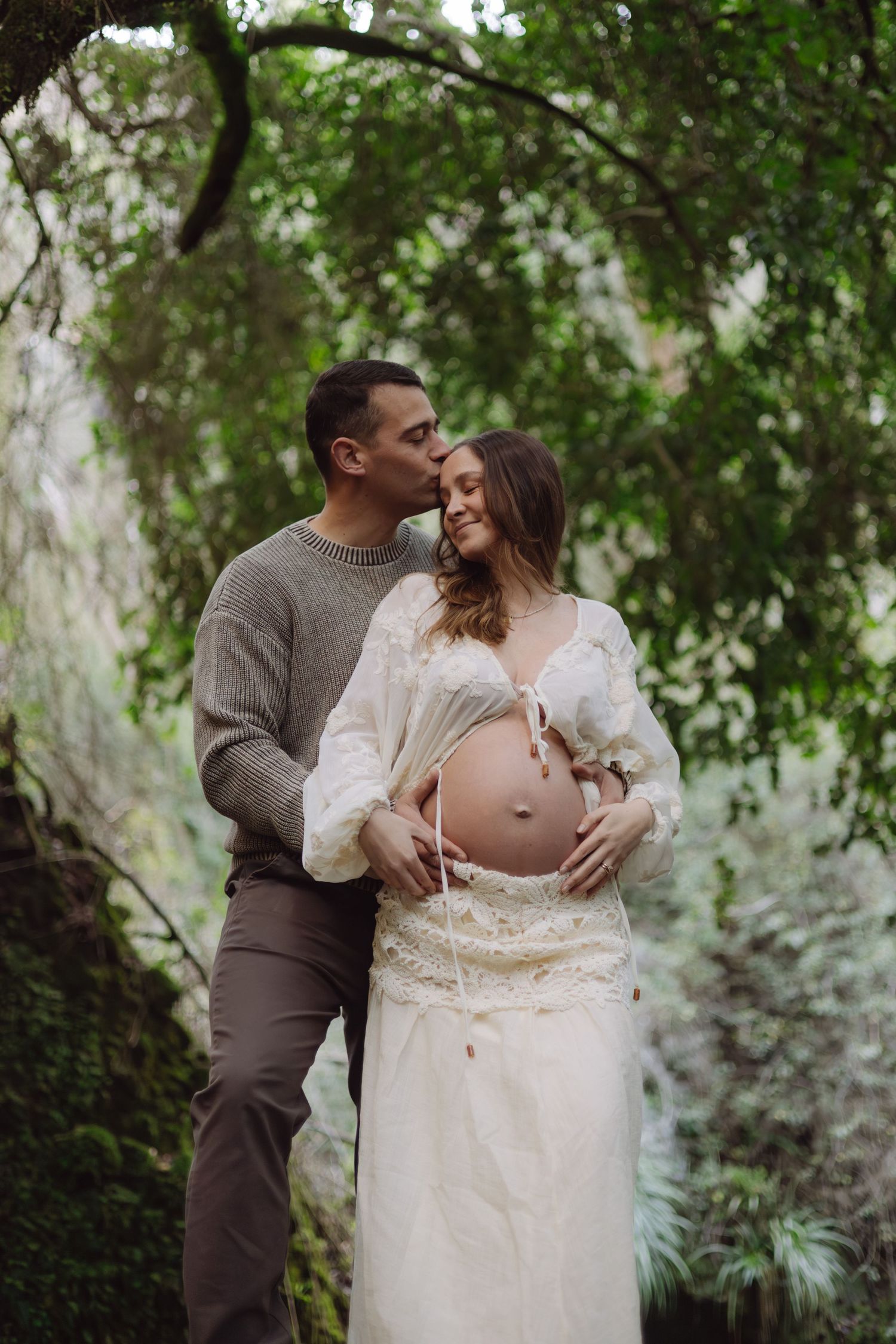A couple poses outdoors, man kissing pregnant woman's head while cradling her baby bump, surrounded by lush green foliage.