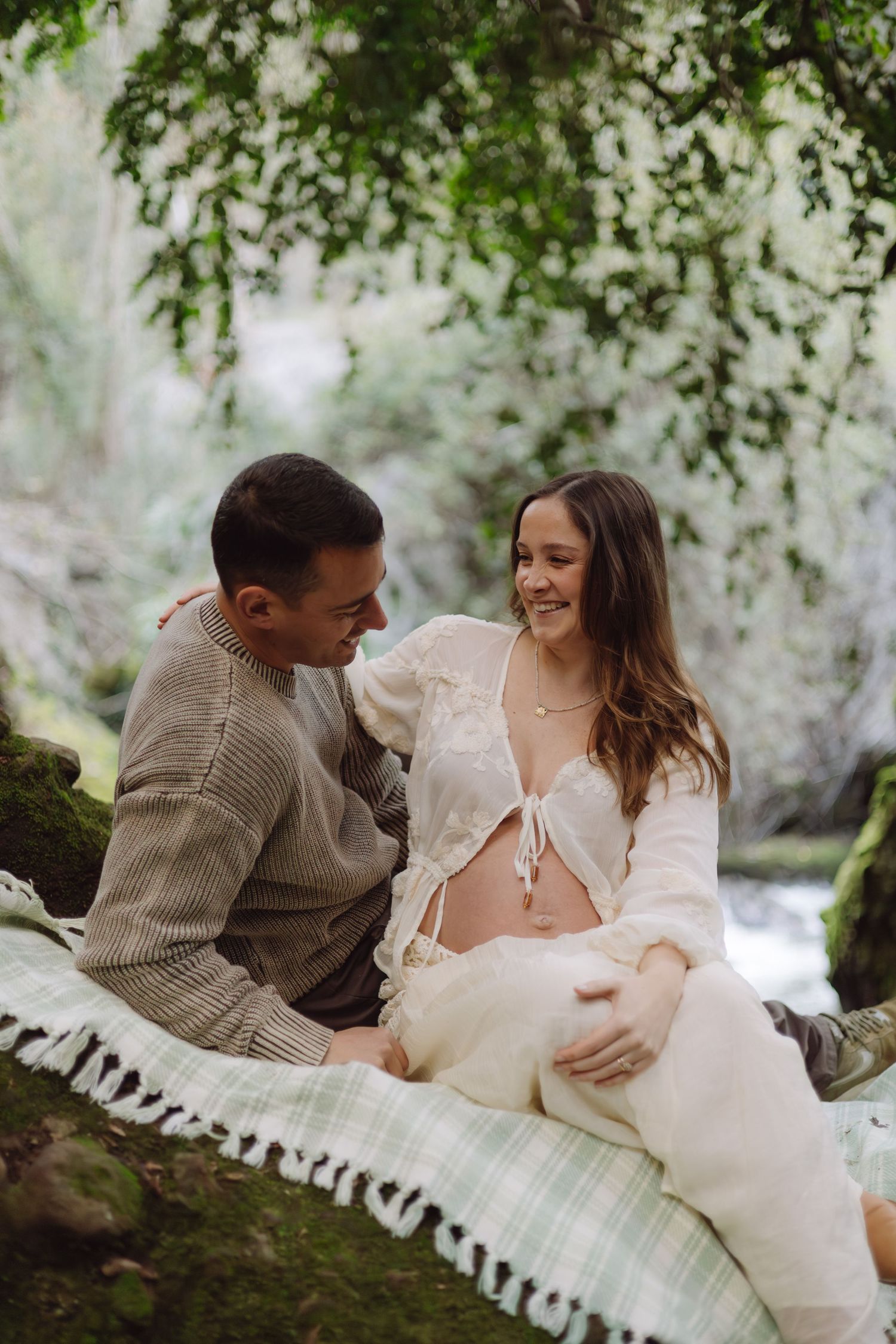 Couple sharing a tender moment during outdoor maternity photoshoot on a blanket in a lush green forest setting.