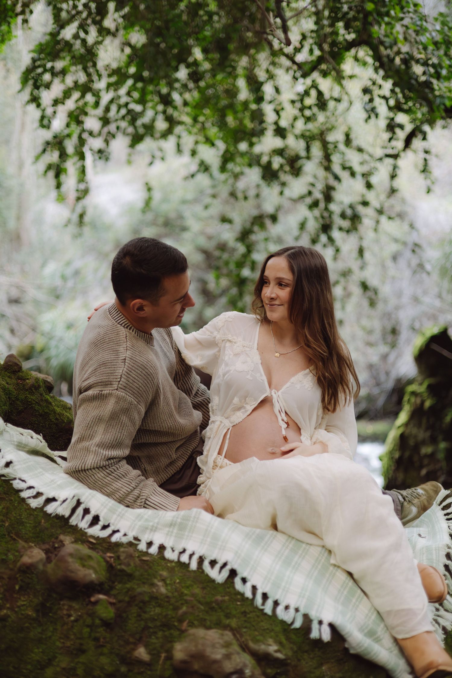 Couple sitting on blanket outdoors, pregnant woman in white lace top smiling at partner near mossy rocks and trees.