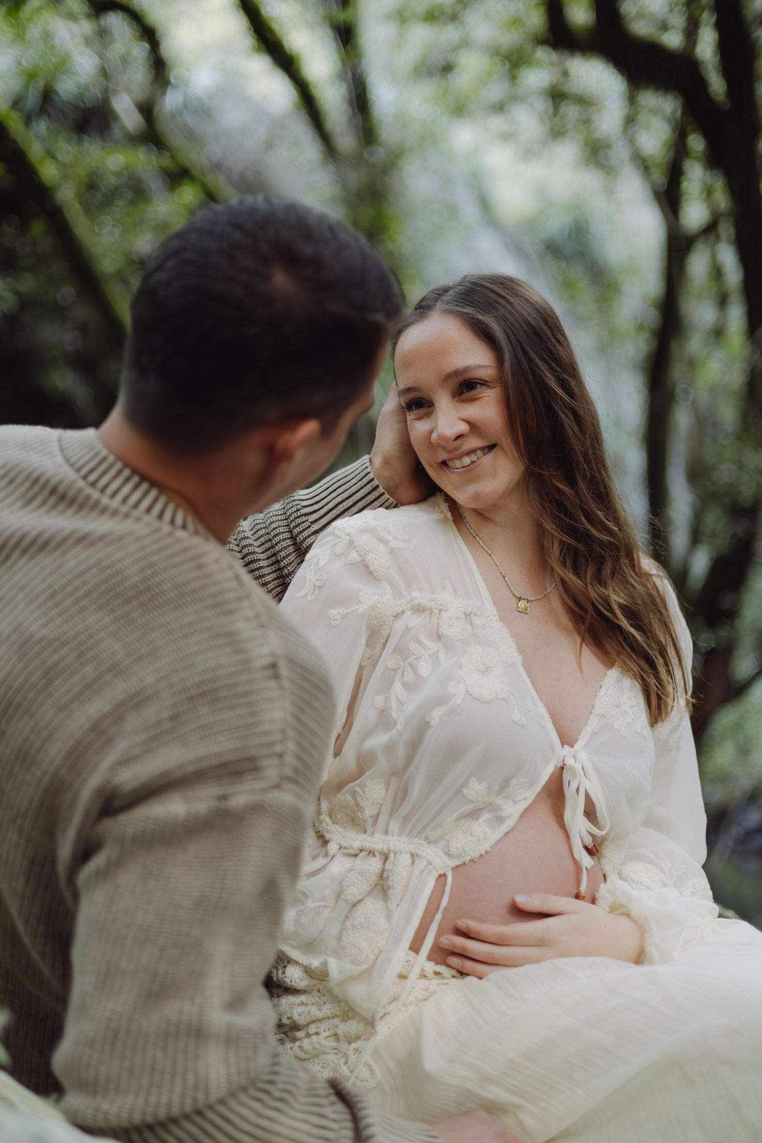 Smiling pregnant woman in white lace dress sits with partner outdoors in lush green forest setting.