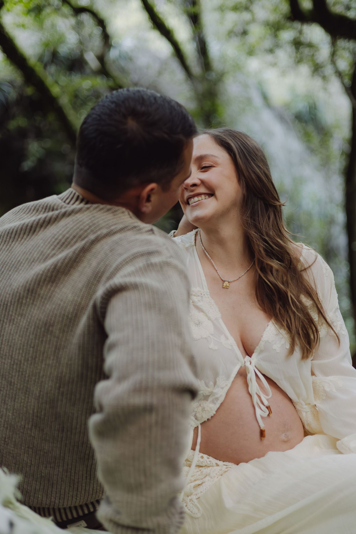Happy pregnant couple sharing intimate moment outdoors, woman in white dress laughing with partner in nature.