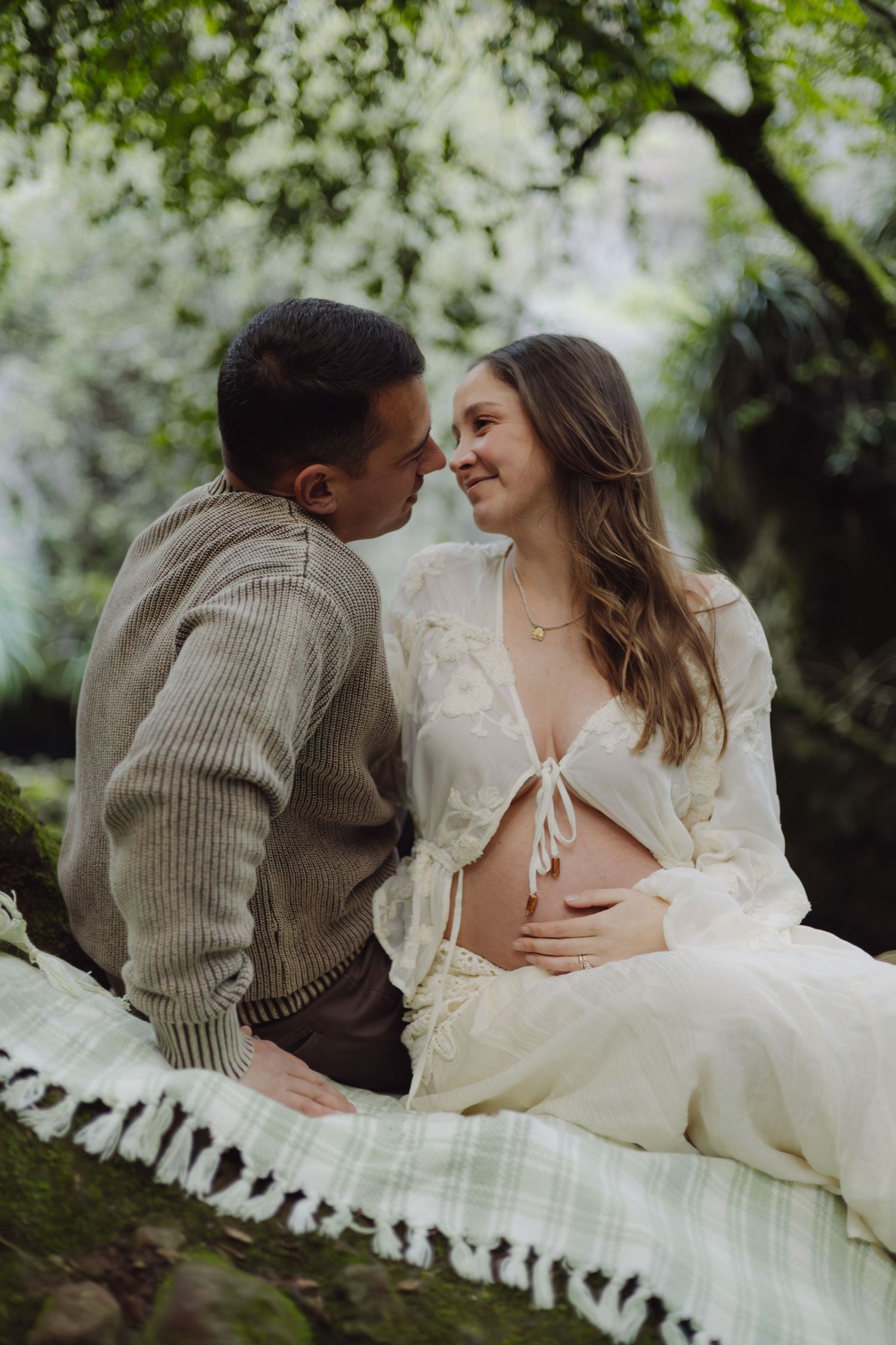 Couple sharing tender moment on blanket outdoors, pregnant woman in white lace dress with exposed belly, surrounded by lush greenery.