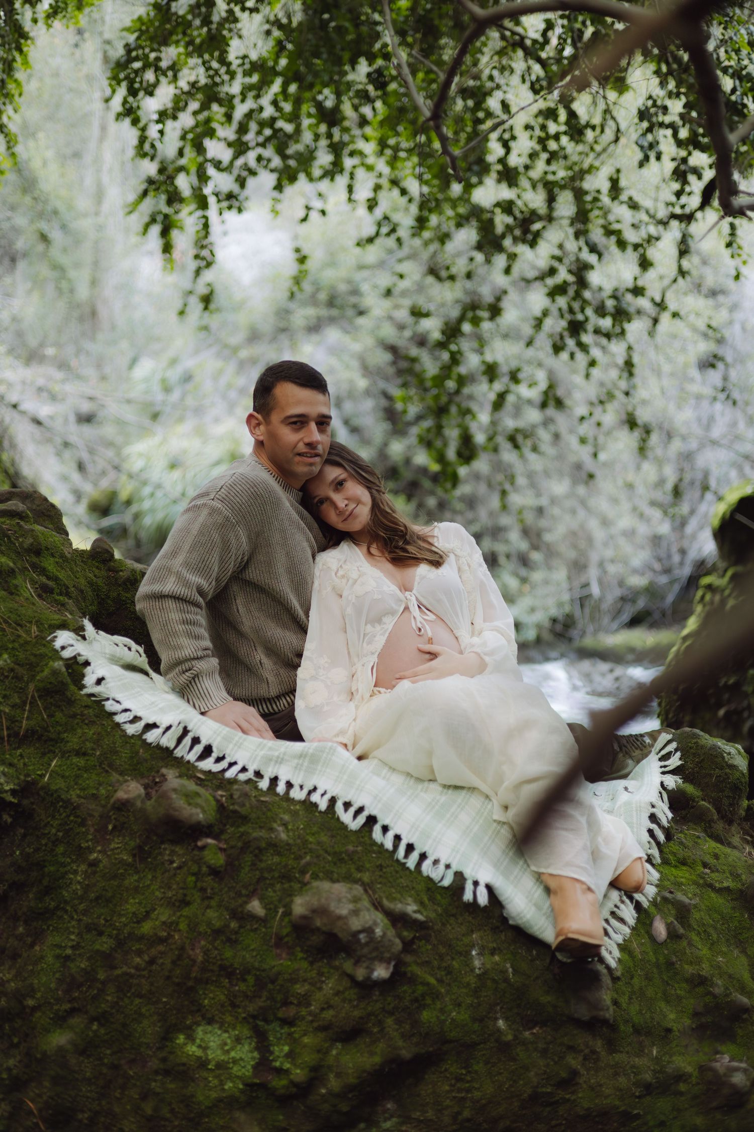 Couple sitting on mossy rocks by a stream, pregnant woman in white dress leaning on partner in gray sweater.