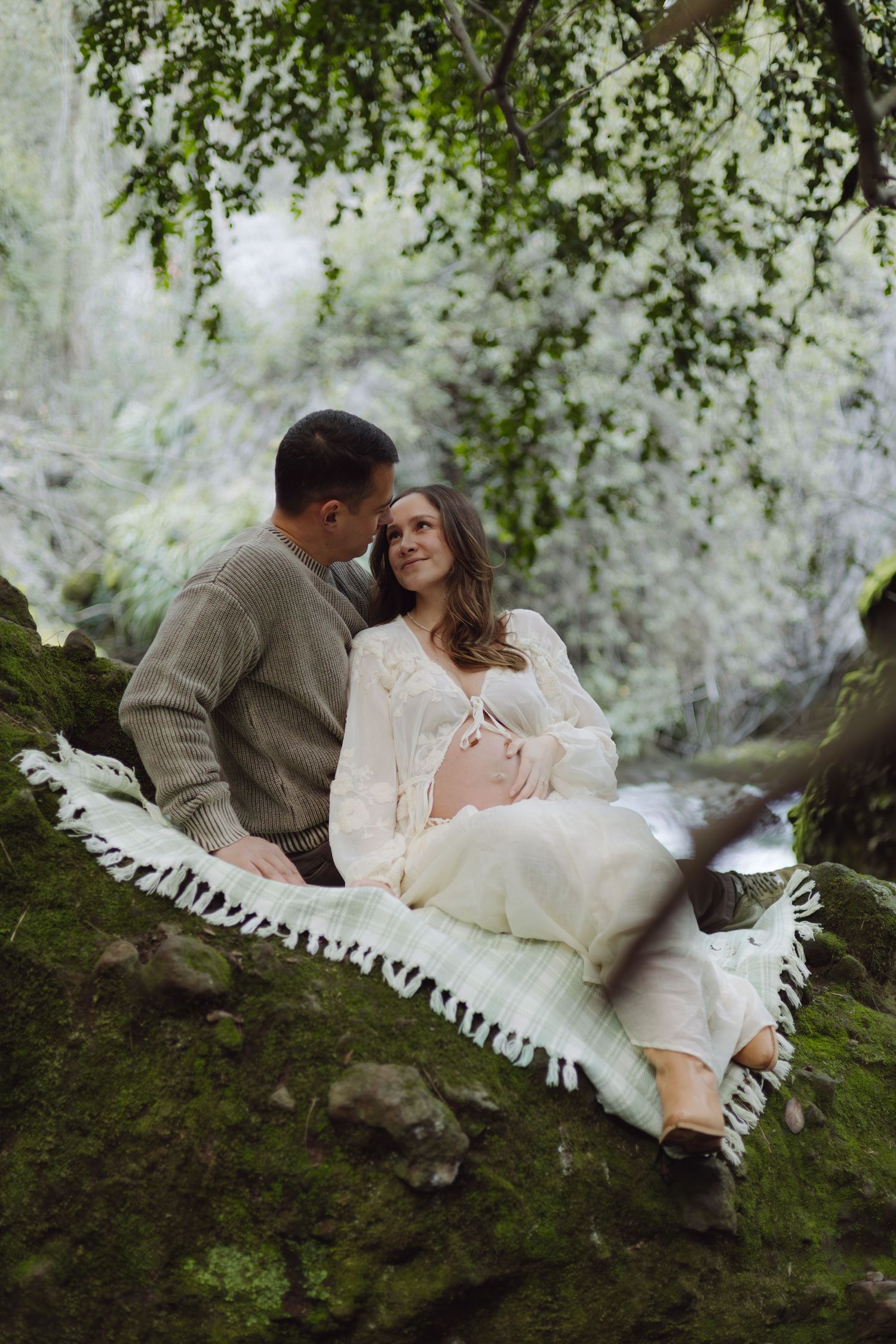 Couple sitting on mossy rocks in a lush forest, pregnant woman in white dress leaning against partner.